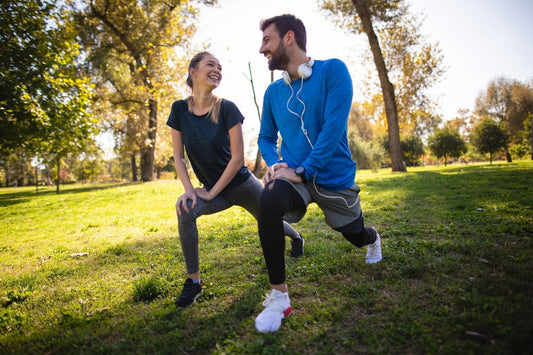 Mann und Frau in Sportkleidung lächeln sich an, während sie Dehnübungen im Park machen bei sonnigem Wetter.