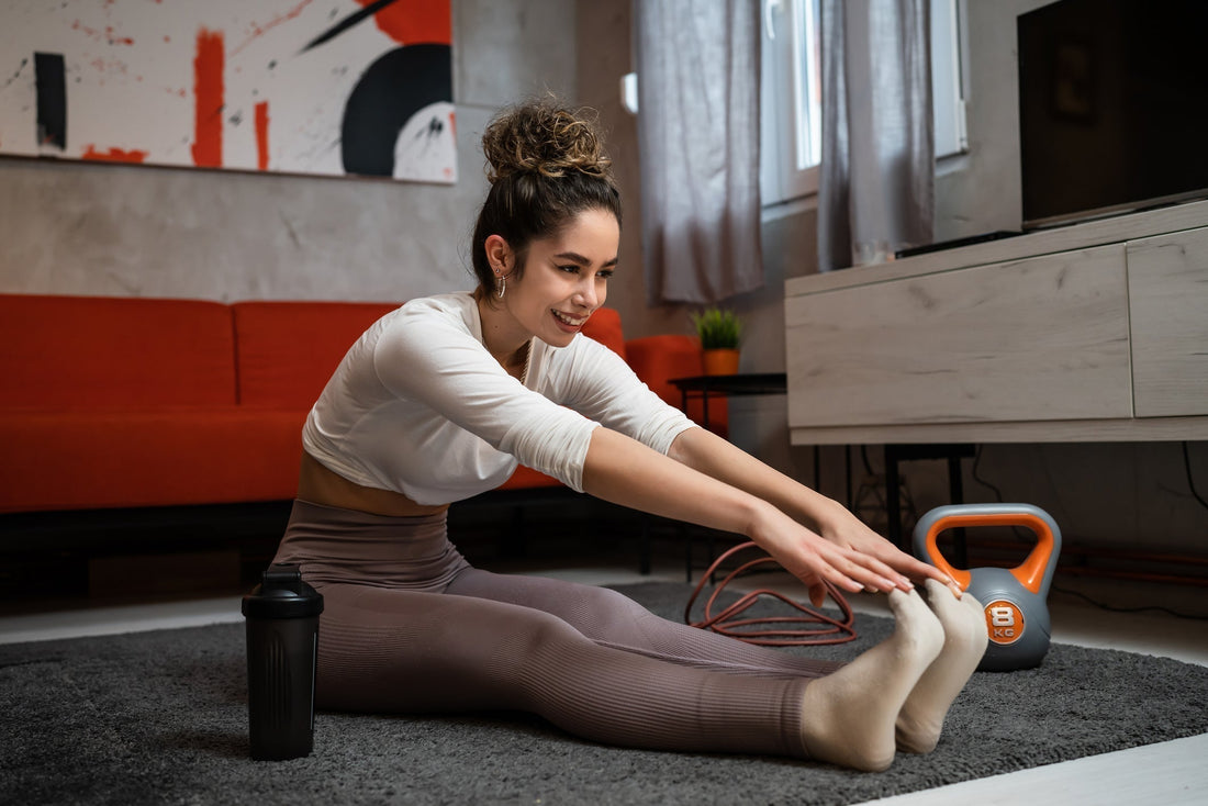 Person sitzt auf einer Trainingsmatte im Wohnraum und beugt den Oberkörper nach vorn zu den ausgestreckten Beinen, Sportequipment liegt daneben. 